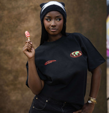 A woman wearing a black graphic t-shirt and a black-and-white striped beanie, holding a red and white striped lollipop against a textured brown wall background.