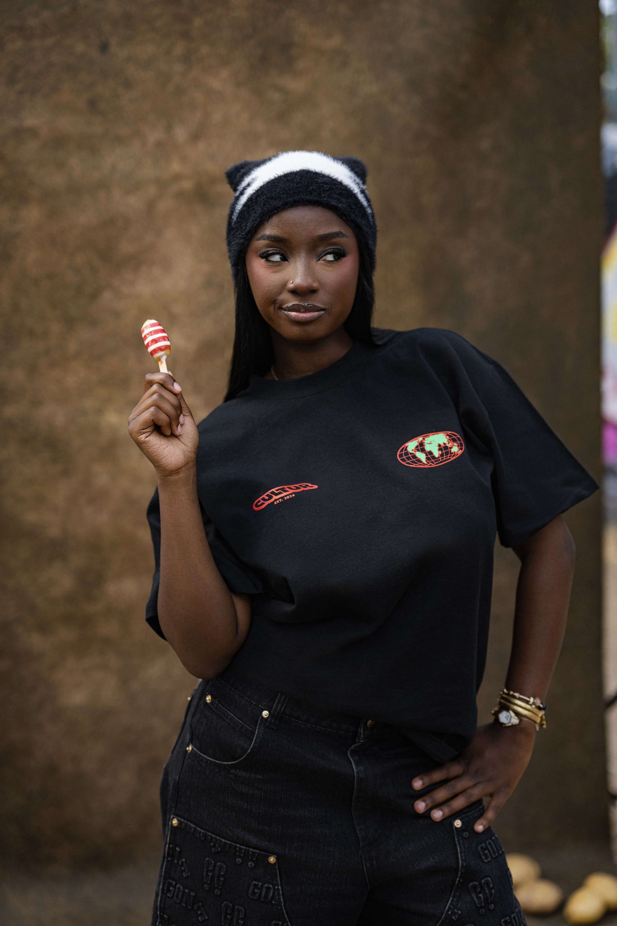 A woman wearing a black graphic t-shirt and a black-and-white striped beanie, holding a red and white striped lollipop against a textured brown wall background.