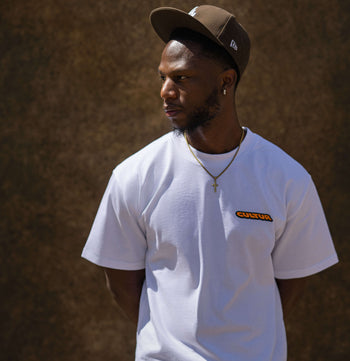 A man wearing a white short-sleeve t-shirt with a small orange and black 'CANTON' logo on the left chest, paired with a brown baseball cap and a gold cross necklace, standing against a textured brown wall.