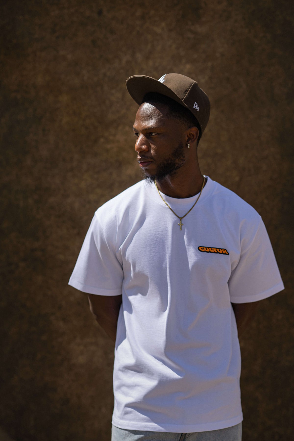 A man wearing a white short-sleeve t-shirt with a small orange and black 'CANTON' logo on the left chest, paired with a brown baseball cap and a gold cross necklace, standing against a textured brown wall.