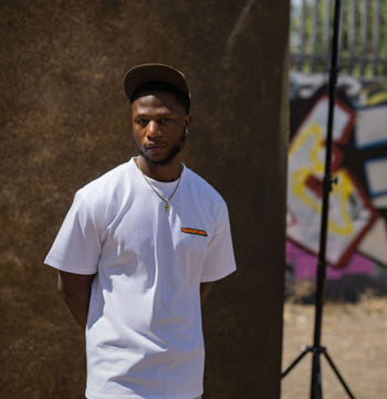 A man wearing a white crewneck t-shirt with a small orange and yellow graphic on the left chest, paired with light blue jeans and a dark cap, standing in front of a brown backdrop in an outdoor setting.