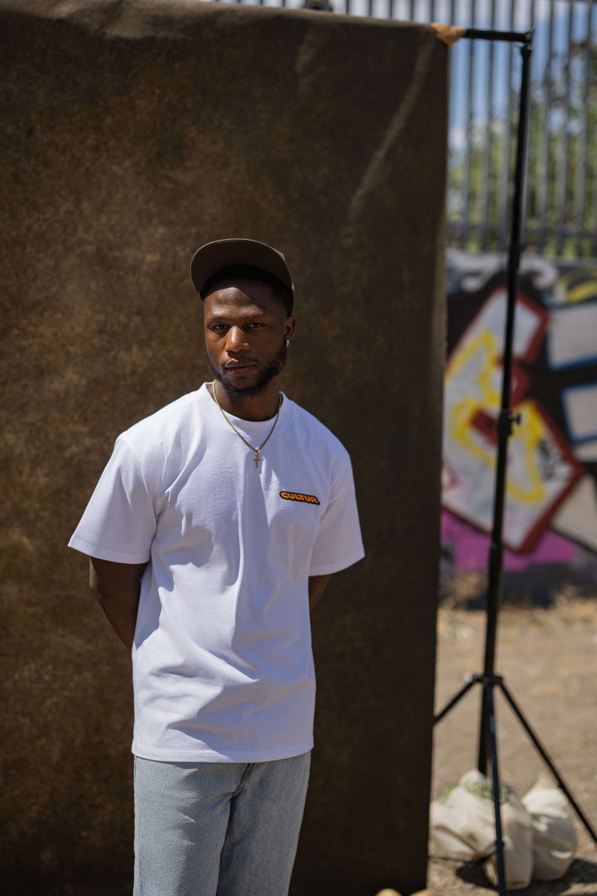 A man wearing a white crewneck t-shirt with a small orange and yellow graphic on the left chest, paired with light blue jeans and a dark cap, standing in front of a brown backdrop in an outdoor setting.