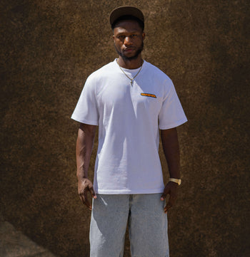 A man wearing a white short-sleeve t-shirt with a small orange and yellow graphic on the left chest, paired with light gray denim shorts and a dark cap, standing against a textured brown wall.