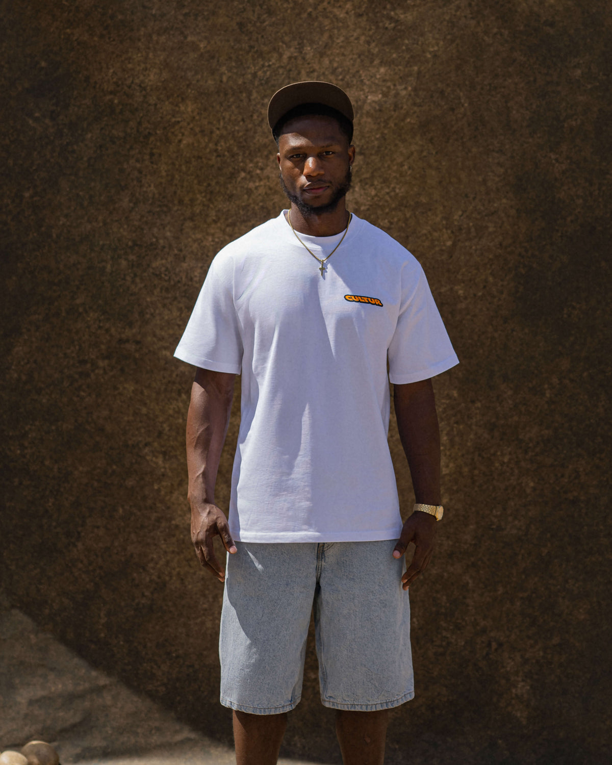 A man wearing a white short-sleeve t-shirt with a small orange and yellow graphic on the left chest, paired with light gray denim shorts and a dark cap, standing against a textured brown wall.