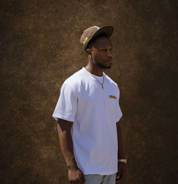 A man wearing a white crewneck t-shirt with a small orange and yellow graphic on the left chest, paired with light blue jeans and a brown baseball cap, standing against a textured brown wall.
