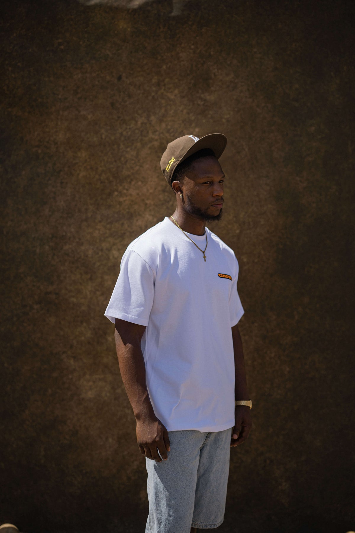 A man wearing a white crewneck t-shirt with a small orange and yellow graphic on the left chest, paired with light blue jeans and a brown baseball cap, standing against a textured brown wall.