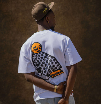 A man wearing a white t-shirt with a large black and orange owl graphic on the back, paired with a brown cap and light blue jeans, standing against a textured brown wall.
