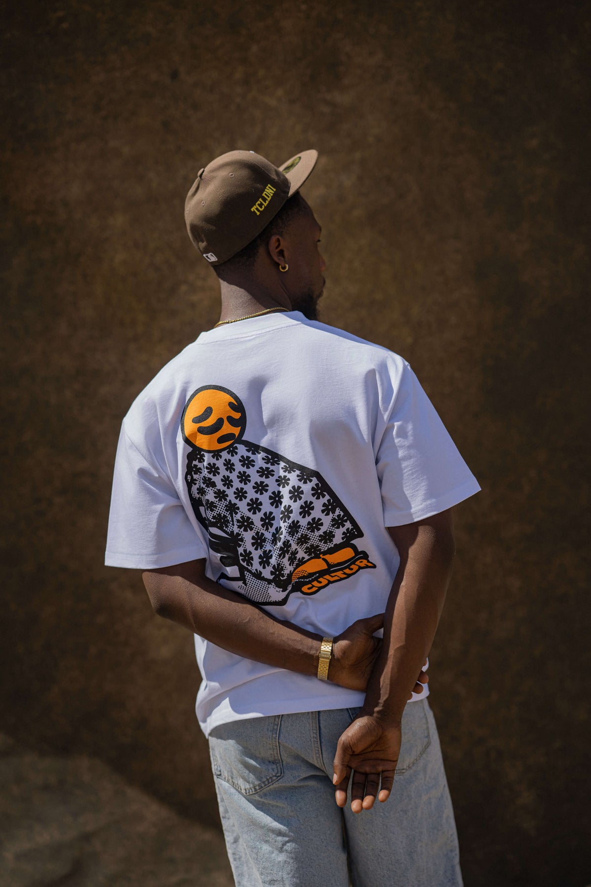 A man wearing a white t-shirt with a large black and orange owl graphic on the back, paired with a brown cap and light blue jeans, standing against a textured brown wall.