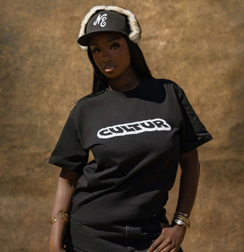 A woman wearing a black t-shirt with a white 'CULTUR' logo across the chest, paired with a black and white striped beanie, standing against a textured brown background.