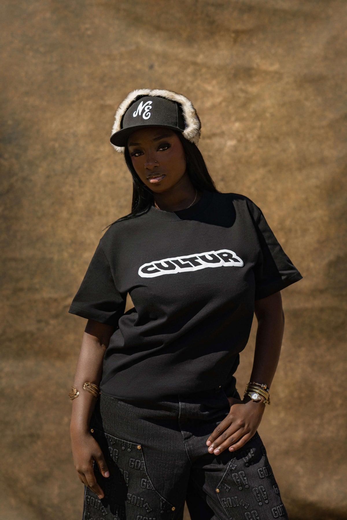 A woman wearing a black t-shirt with a white 'CULTUR' logo across the chest, paired with a black and white striped beanie, standing against a textured brown background.